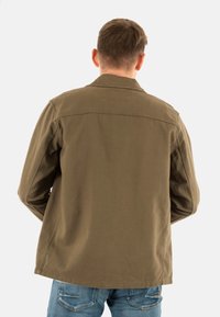 Veste en toile vert olive avec un col pointu, des manches longues et une coupe droite, associée à un jean en denim bleu.