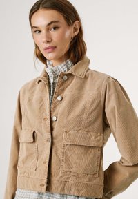 Beige corduroy jas met zilveren drukknoopjes, twee voorzakken en een knoopsluiting. Gelaagd over een geruit shirt met een hoge kraag.