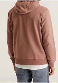 Hombre con una sudadera color rosa viejo sobre una camiseta blanca y jeans azules, visto desde atrás contra un fondo claro y liso.