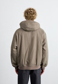 Přes oversized béžová bunda s kapucí, která má elastické manžety a pás. Hladká textura látky, jednoduchý design s prošitými detaily.