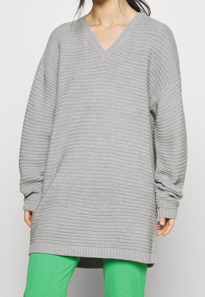 Maglione grigio lavorato a maglia, oversize, con scollatura a V, polsini a costina e un motivo testurizzato. Abbinato a leggings verdi a coste.