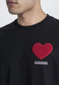 Černé tričko s texturovanou červenou nášivkou srdce na levé straně a bílým vyšitým textem "KAVANAGH" pod nášivkou.