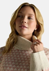 Junge Frau mit blonden Haaren, die einen cremefarbenen und rosa gemusterten Rollkragenpullover, einen silbernen Ring und silberne Ohrringe trägt, schaut leicht zur Seite.