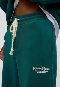 Groene sweatpants van zachte stof met een trekkoord in de taille. Bevat een zijzak en geborduurde tekst op de dij in het wit.