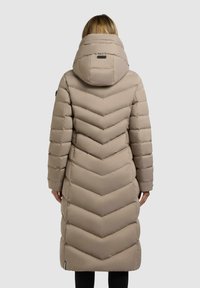 Donna che indossa un lungo cappotto trapuntato beige con un ampio cappuccio, vista da dietro su uno sfondo neutro.