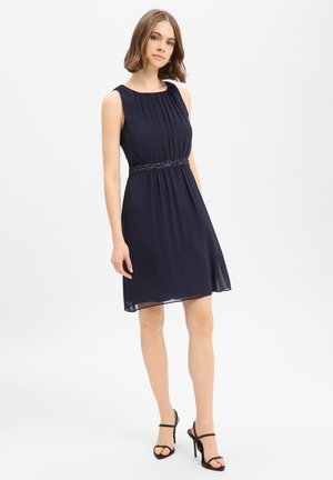 Jonge vrouw met een mouwloze marineblauwe knieëlange jurk met een tailleband met kralen en zwarte sandalen met hoge hakken, staand tegen een witte achtergrond.