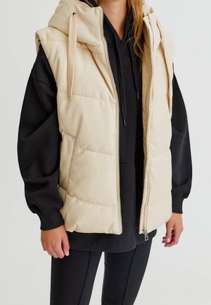 Person trägt eine beige ärmellose gesteppte Weste mit Kapuze über einem schwarzen Hoodie mit langen Ärmeln und schwarzen Hosen, steht mit entspannten Armen.