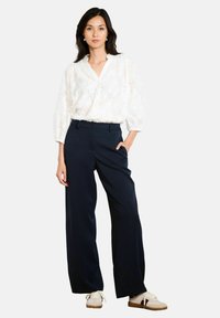 Blusa bianca in pizzo floreale con vestibilità ampia e scollatura a V, abbinata a pantaloni a gamba larga blu navy con vita alta e sneaker beige.