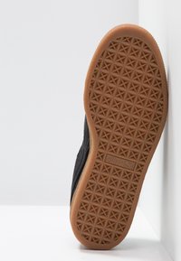 Semelle d'une chaussure Puma noire avec une bande de roulement en caoutchouc brun texturé présentant des motifs géométriques et le logo Puma au centre.