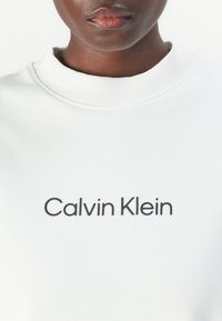 Biela mikina vyrobená z bavlny, s rebrovaným výstrihom a tučným čiernym textom "Calvin Klein" na hrudi. Jednoduchý, čistý dizajn.