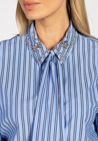 Vrouw in een lichtblauw gestreept bloesje met een strik en rhinestone versieringen op de kraag.
