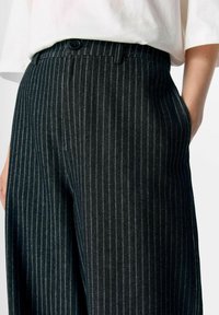 Schwarz gestreifte Hose mit taillierter Passform, ausgestattet mit einem Knopfverschluss und schrägen Seitentaschen. Hergestellt aus strukturiertem Stoff.