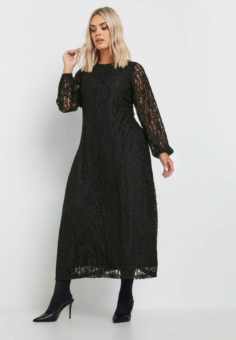 Svart maxiklänning i spets med långa puffärmar och rund halsringning. Har blommönster och är kombinerad med svarta klackar och strumpbyxor.