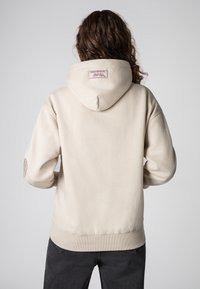 Beige hoodie met een grote capuchon aan de achterkant, voorzien van een label met tekst. Geribbelde manchetten en zoom, gladde stofstructuur. Zichtbare stikseldetails.