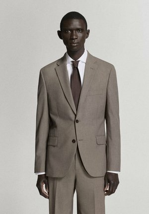 Hombre vestido con un traje beige, camisa blanca y corbata marrón, de pie frente a un fondo claro y liso mirando hacia adelante.