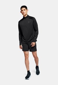 Haut de sport noir à manches longues avec zip quart et logo, associé à un short noir, porté avec des chaussures de sport noires et grises.