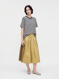 Femme debout portant un t-shirt rayé noir et blanc à manches courtes, une jupe midi plissée jaune moutarde et des chaussures plates beiges.
