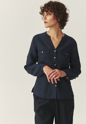 Kobieta o krótkich kręconych włosach, ubrana w ciemnoniebieską koszulę na guziki z kieszeniami na piersiach oraz czarne spodnie, patrząca na bok, trzymająca ręce złożone razem.