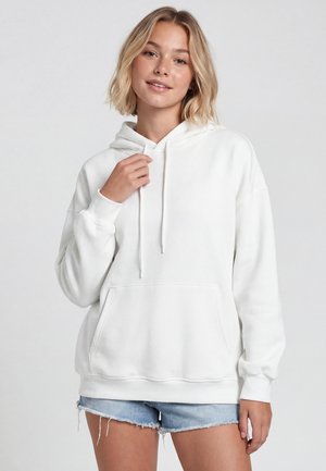 Tânără cu păr blond scurt, purtând un hanorac alb și pantaloni scurți din denim albastru, stând în fața unui fundal simplu și luminos.