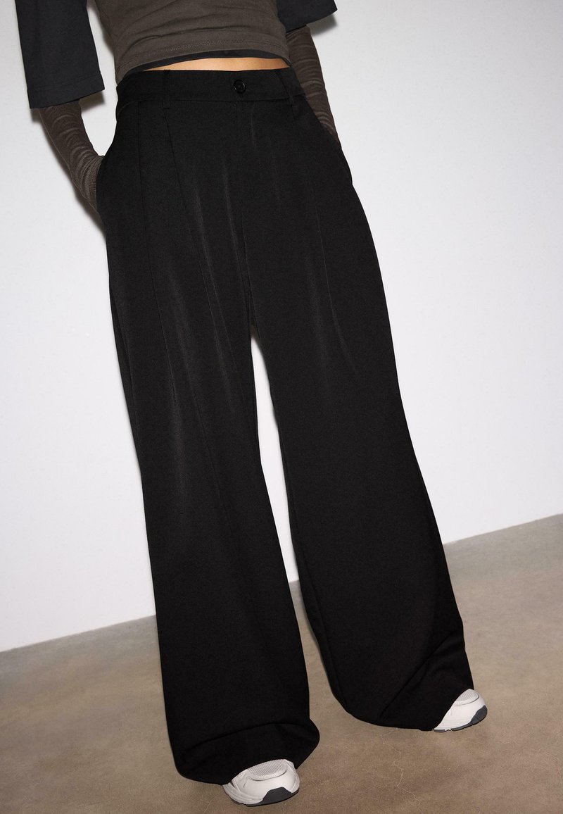 Personne portant un pantalon noir taille haute à jambes larges, des hauts à manches longues superposés foncés, et des baskets blanches, debout devant un fond clair uni.