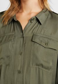 Camicia corta a maniche corte verde oliva con due tasche sul petto, chiusura con bottoni e una texture liscia e leggermente lucida. Presenta un colletto classico.