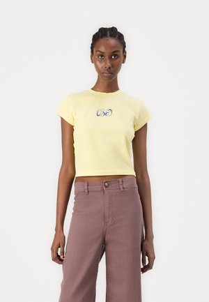 Un t-shirt court en coton jaune avec des manches courtes, présentant "OBEY" en lettres bleues. Associé à un pantalon mauve taille haute et large.