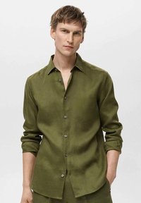 Camicia verde oliva con bottoni, maniche lunghe e colletto appuntito, realizzata in un tessuto testurizzato, con bottoni frontali e polsini arrotolati.