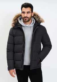 Giacca a vento nera con cappuccio rifinito in pelliccia, dotata di chiusura con zip, texture trapuntata e tasche laterali sopra un hoodie grigio.