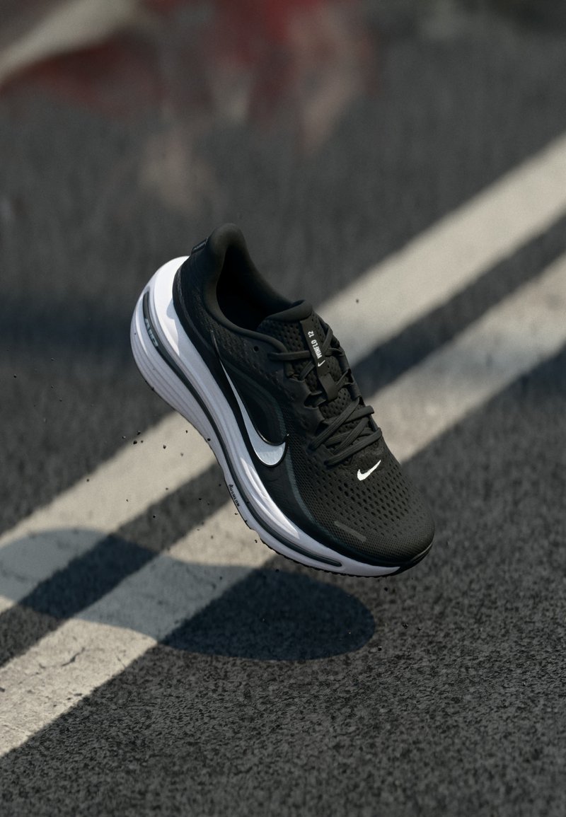 Chaussure de course Nike noire et blanche en l'air au-dessus d'une route en asphalte avec des marquages blancs au sol.