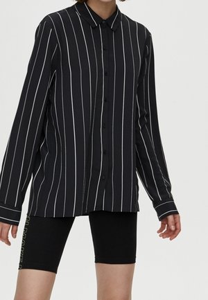 Chemise noire à manches longues boutonnée avec des rayures blanches verticales portée avec un short noir ajusté.