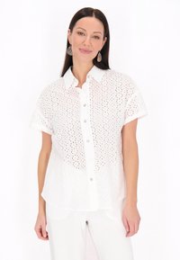 Camicetta bianca a maniche corte con bottoni realizzata in tessuto perforato, con colletto classico e orlo dritto, abbinata a pantaloni bianchi.