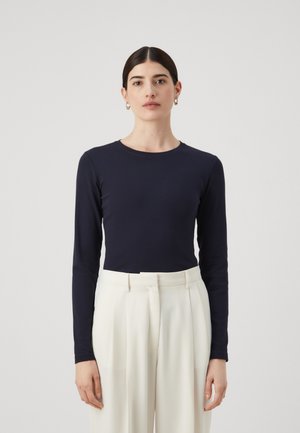 Marineblå langermet topp med rund hals, kombinert med hvite bukser med høy midje. Glatt tekstur, tettsittende design. Gyldne hoop øredobber synlige.