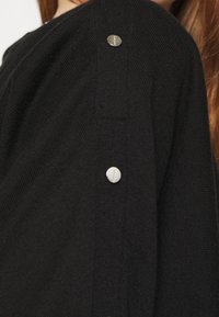 Sort striktrøje med et tekstureret design og sølvknapdetaljer på skuldrene. Blødt stof med en afslappet pasform, der fremhæver fine detaljer.