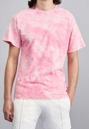Osoba ubrana w różowo-biały krótki rękaw Nike z efektem tie-dye i białe spodnie na jednolitym szarym tle.