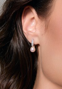 Kolczyki z perłami w kształcie kropli, z perłą w kształcie łzy i srebrnym akcentem z małymi przezroczystymi kamieniami, na tle ciemnych włosów.