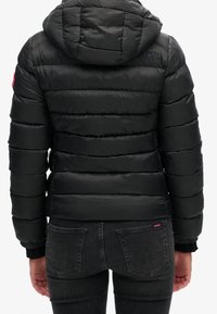 Persona che indossa un giubbotto imbottito nero a quadratini con cappuccio e jeans scuri, vista da dietro su uno sfondo semplice.
