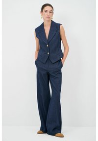Tmavě modrá tailored vesta s klopovým límcem a předními knoflíky, spárovaná se širokými kalhotami ze stejné látky, které mají boční kapsy.