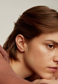 Pendiente de aro dorado con una textura sutil, llevado en la oreja. Vista en primer plano de la cara de una persona, mostrando una piel suave y cabello corto castaño.