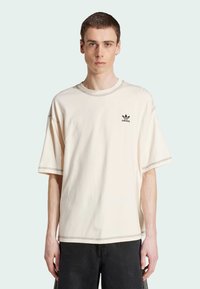 T-shirt oversize in cotone crema, con collo rotondo, maniche corte e logo adidas nero sul lato sinistro del petto; cuciture a contrasto visibili.