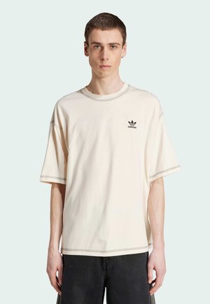 Krem majica širokog kroja od pamuka, s okruglim izrezom, kratkim rukavima i crnim adidas logotipom na lijevoj strani grudi; vidljivo kontrastno šivanje.