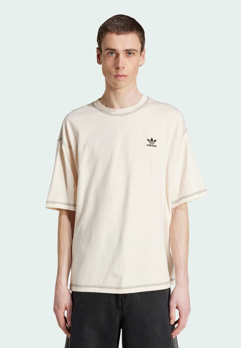 T-shirt oversize in cotone crema, con collo rotondo, maniche corte e logo adidas nero sul lato sinistro del petto; cuciture a contrasto visibili.