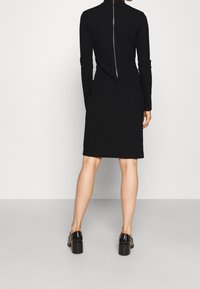 Robe noire à manches longues avec une silhouette ajustée, col haut, fermeture éclair visible au dos et tissu lisse ; portée avec des chaussures à talons épais.