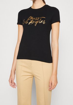 Femme portant un t-shirt ajusté noir avec un texte doré brillant « Guess Los Angeles », associé à un pantalon beige taille haute.