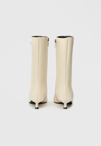 Bottes crème montantes en matériau lisse, avec un bout effilé, un talon évasé et un zip visible à l'arrière.