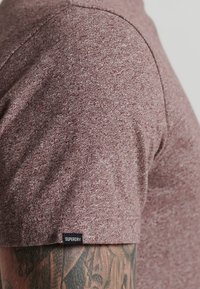 Czerwona, melanżowa, krótko rękawowa koszula wykonana z teksturowanej tkaniny. Posiada mał�ą czarną metkę "Superdry" blisko szwu dolnego. Zbliżenie na okolicę ramienia.