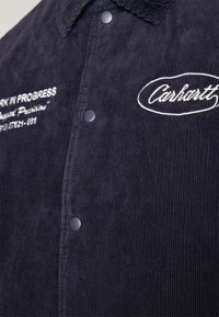 Nahaufnahme eines marineblauen Cordhemds mit Druckknöpfen, das ein weiß gesticktes "Carhartt"-Logo und einen teilweisen Text "Work in Progress" zeigt.