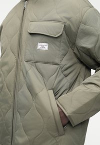 Šviesiai alyvuogių žalios spalvos striukė su siūtais raštais, su užtrauktuku, krūtinės kišenė su užklotėliu ir užrašu „Guess Jeans Authentic“, bei šonine ranka kišene, dėvima su įkišta ranka.