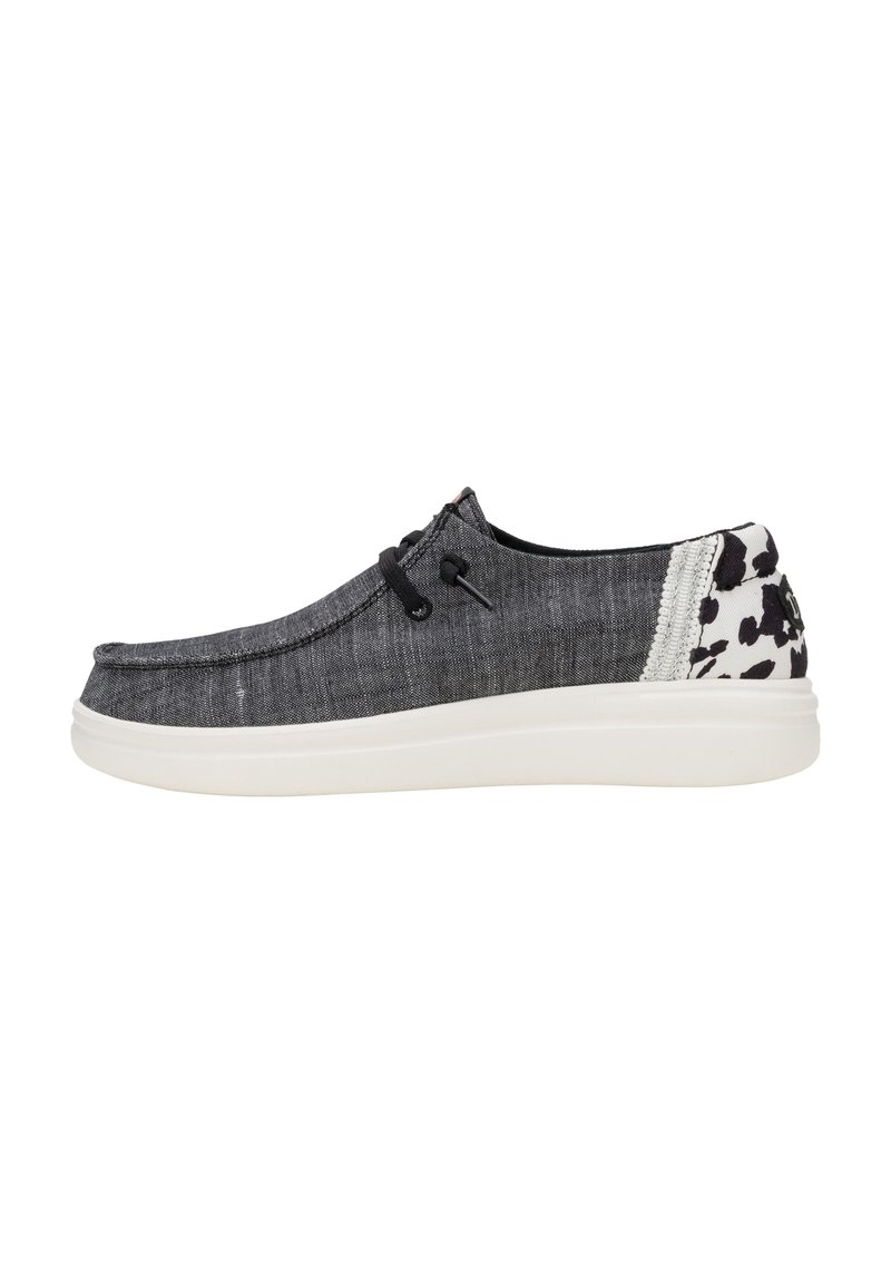 Zapato náutico de mezclilla negra con suela blanca, que presenta cordones negros y un acento estampado en el talón. Textura suave con un diseño casual.