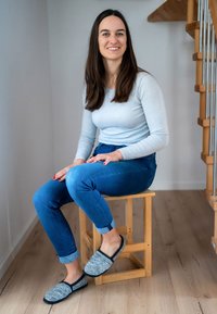 Grijze en zwarte vilten pantoffels hebben een ronde neus en een instapontwerp. De drager zit op een houten kruk en draagt een lichtblauw shirt met lange mouwen en blauwe jeans.
