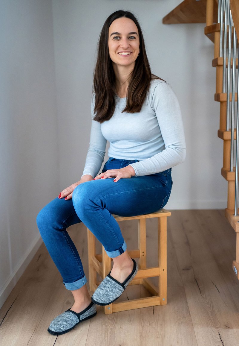 Grijze en zwarte vilten pantoffels hebben een ronde neus en een instapontwerp. De drager zit op een houten kruk en draagt een lichtblauw shirt met lange mouwen en blauwe jeans.